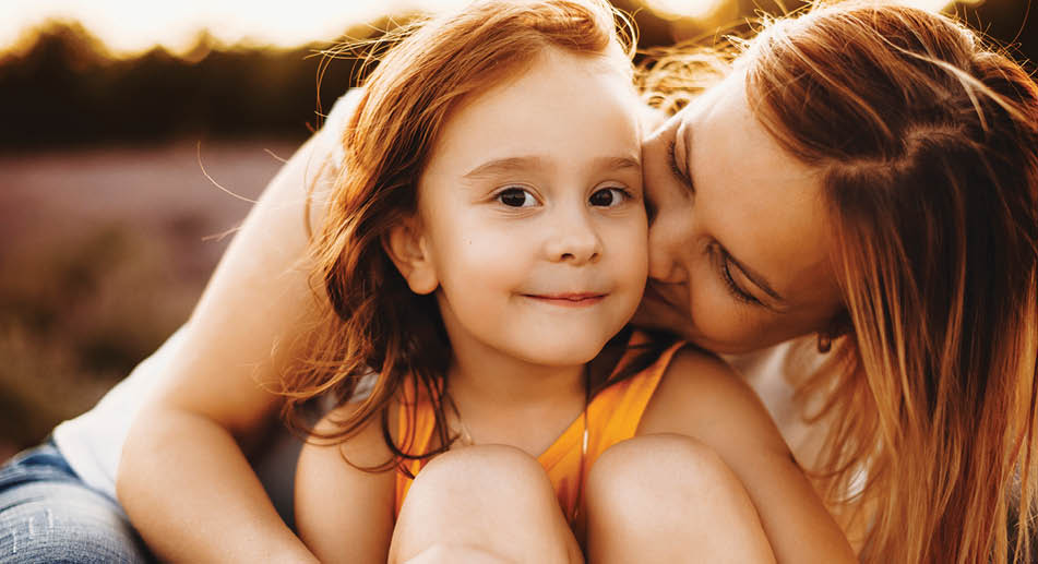 Close up portrait of a amazing little girl looking at camera smiling while being emraced and kised by her mother against sunset.