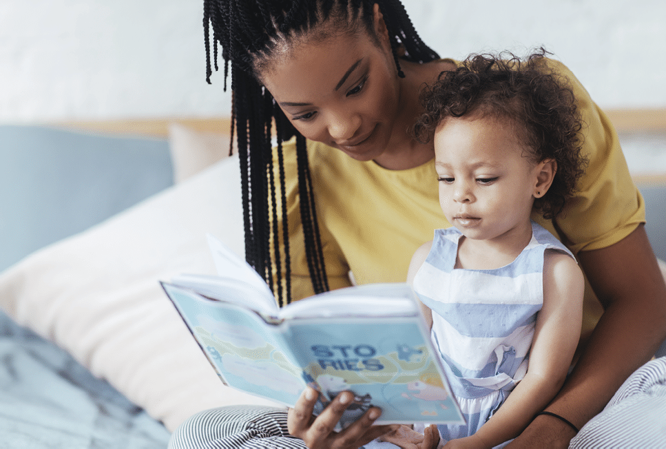 Beautiful African woman sitting on bed and reading a book to her cute baby daughter.