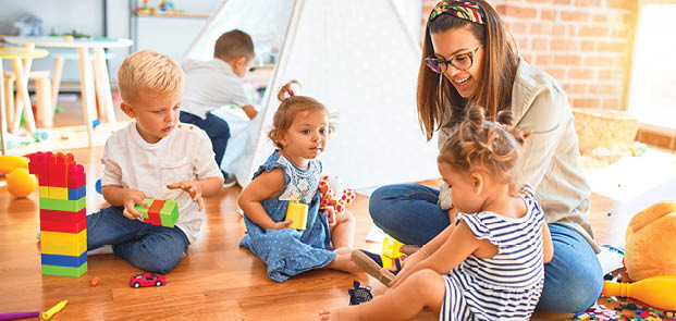Beautiful teacher and group of toddlers playing around lots of toys at kindergarten